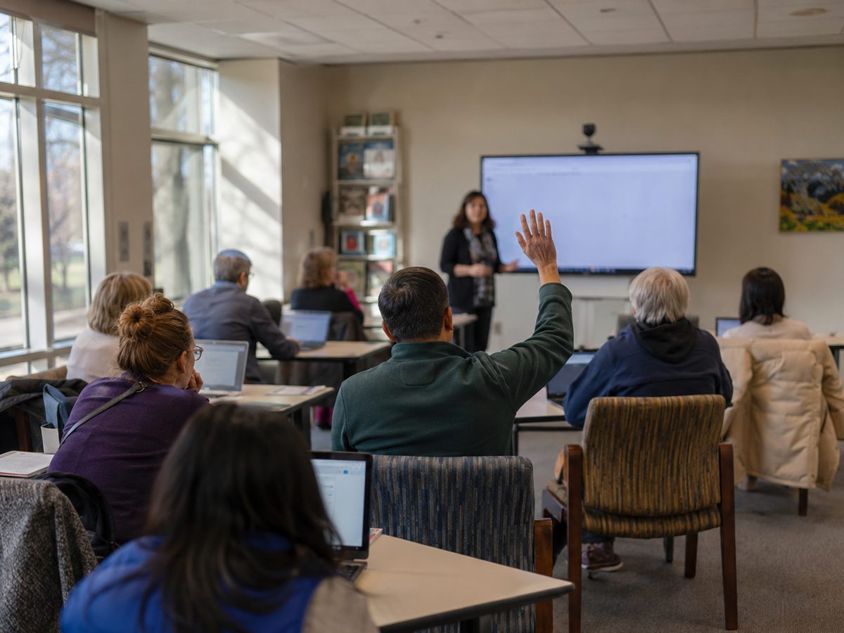 Participants seated at tables with laptops during a Tech Ready Nevada workshop, one raising a hand to ask the instructor a question.